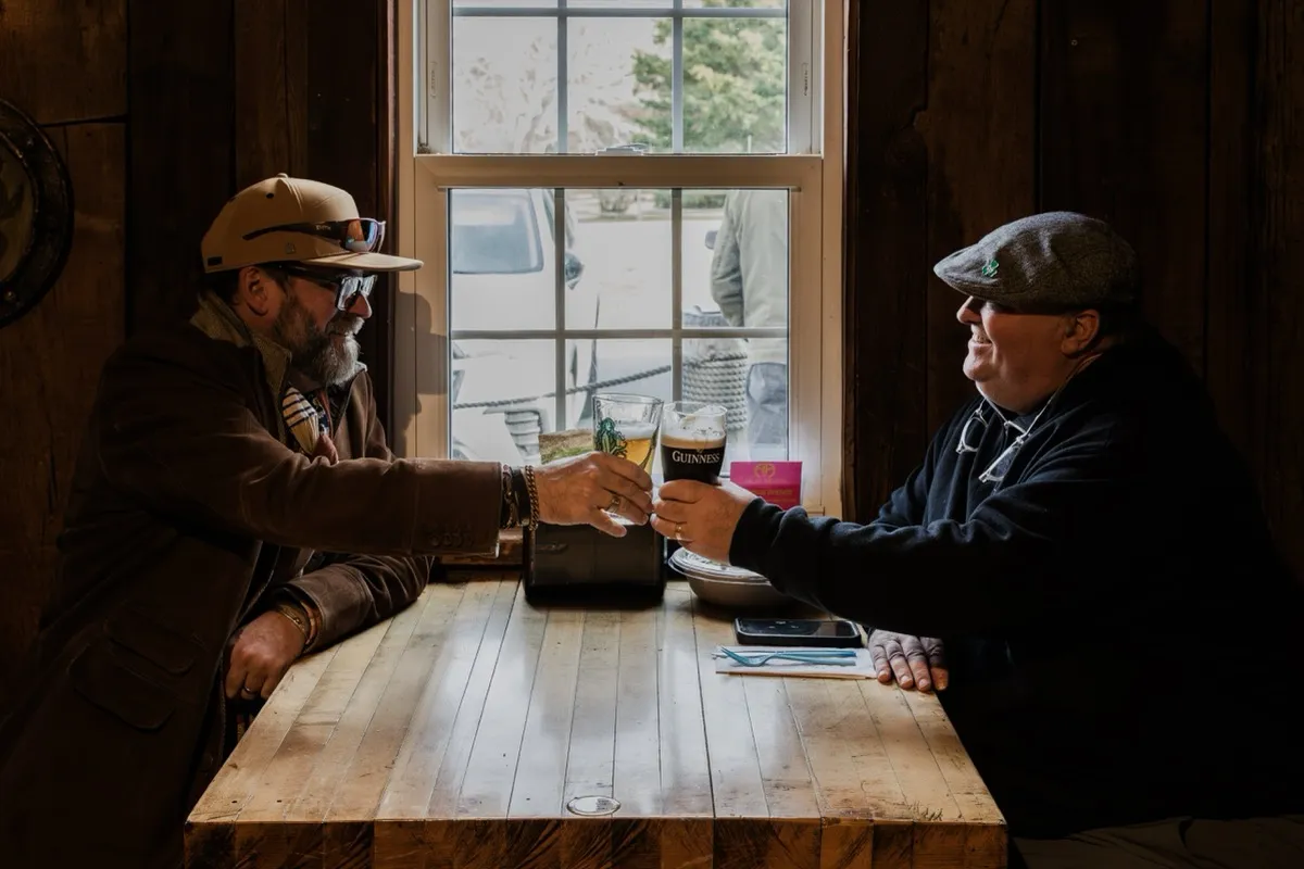 Two regulars raising a pint and a glass at a window table